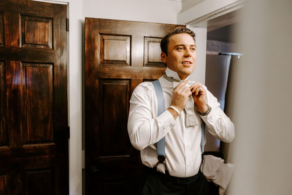 Groom in white shirt and suspenders adjusting his bow tie before wedding ceremony.