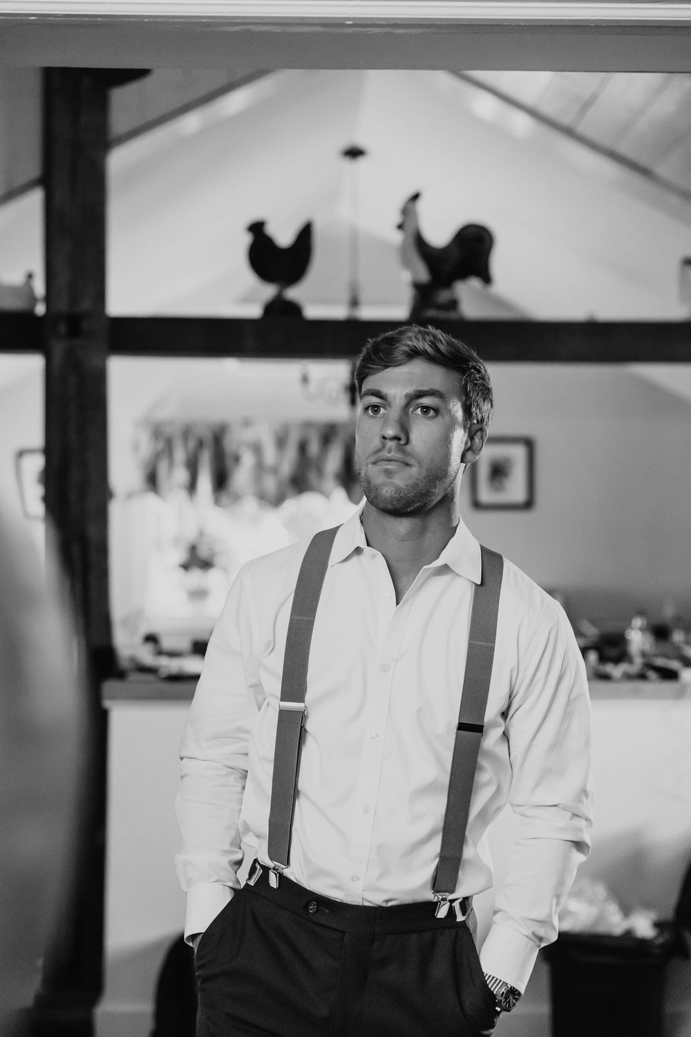 Black and white portrait of groom in white shirt and suspenders standing confidently indoors.