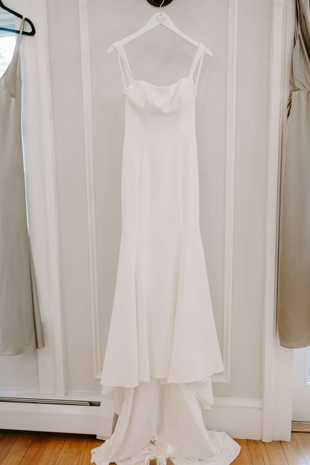 Elegant white spaghetti strap mermaid wedding gown hanging on hanger against white door.