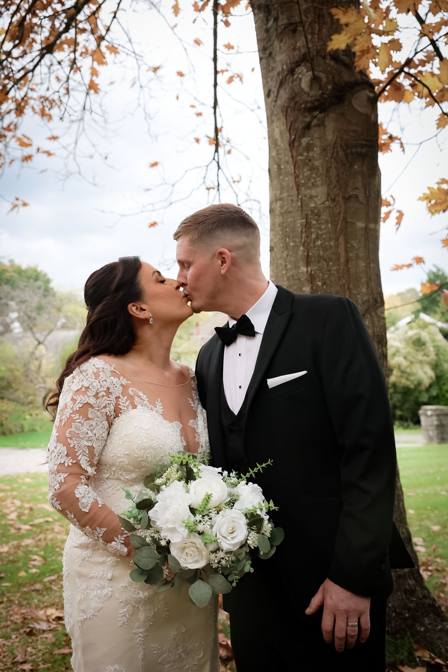 Bride and groom kissing under an autumn tree, bride holding white rose bouquet, groom in classic black tuxedo.