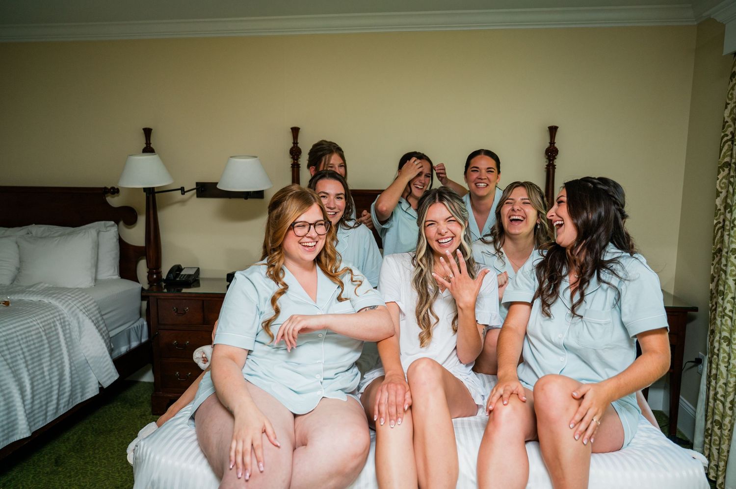 A group of friends in matching light blue and white outfits laugh together while sitting on a bed in a hotel room.