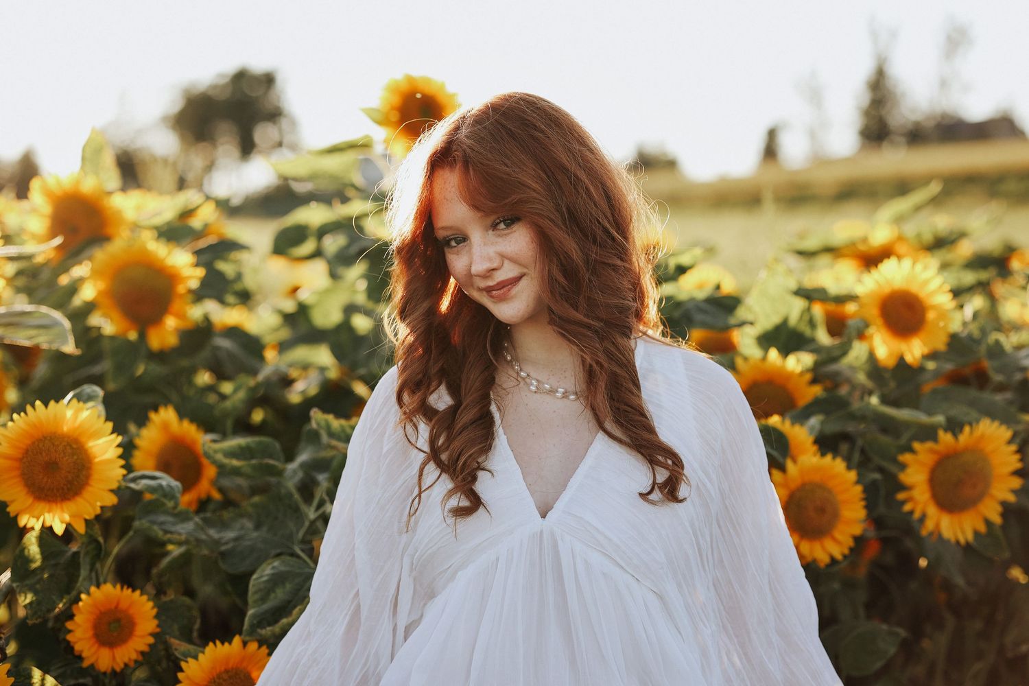 A figure in a white dress stands among vibrant yellow sunflowers in a golden summer field at sunset.