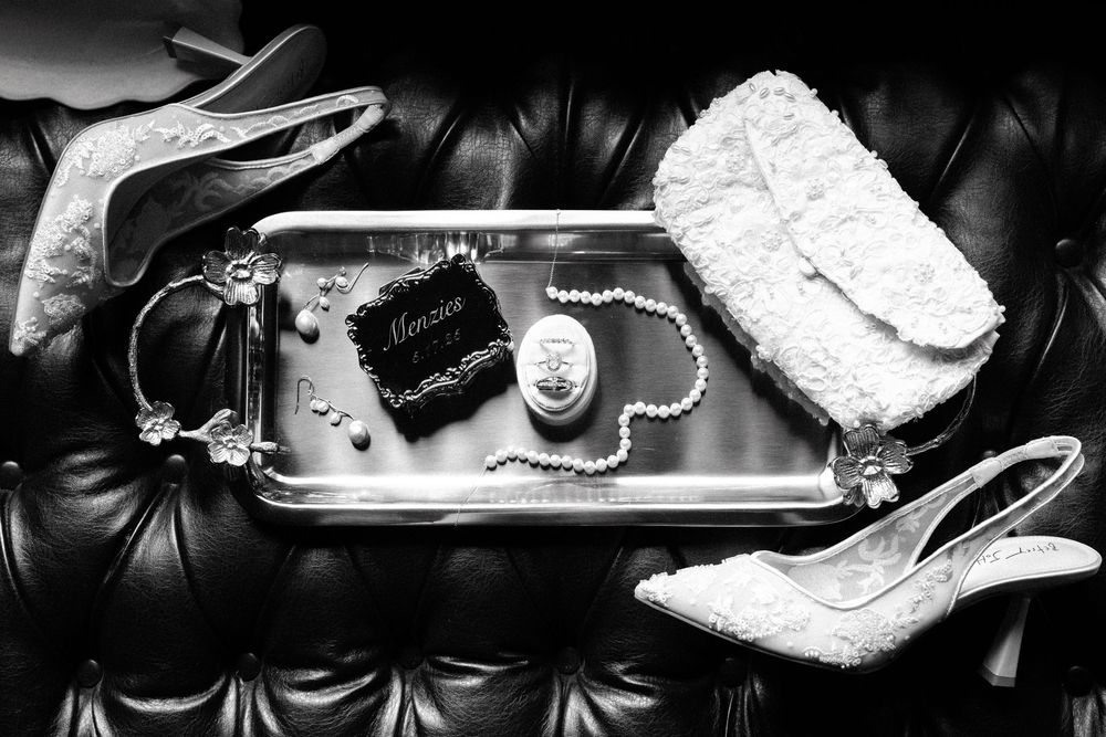 Wedding accessories including white lace heels, jewelry, and a clutch arranged on black leather surface.