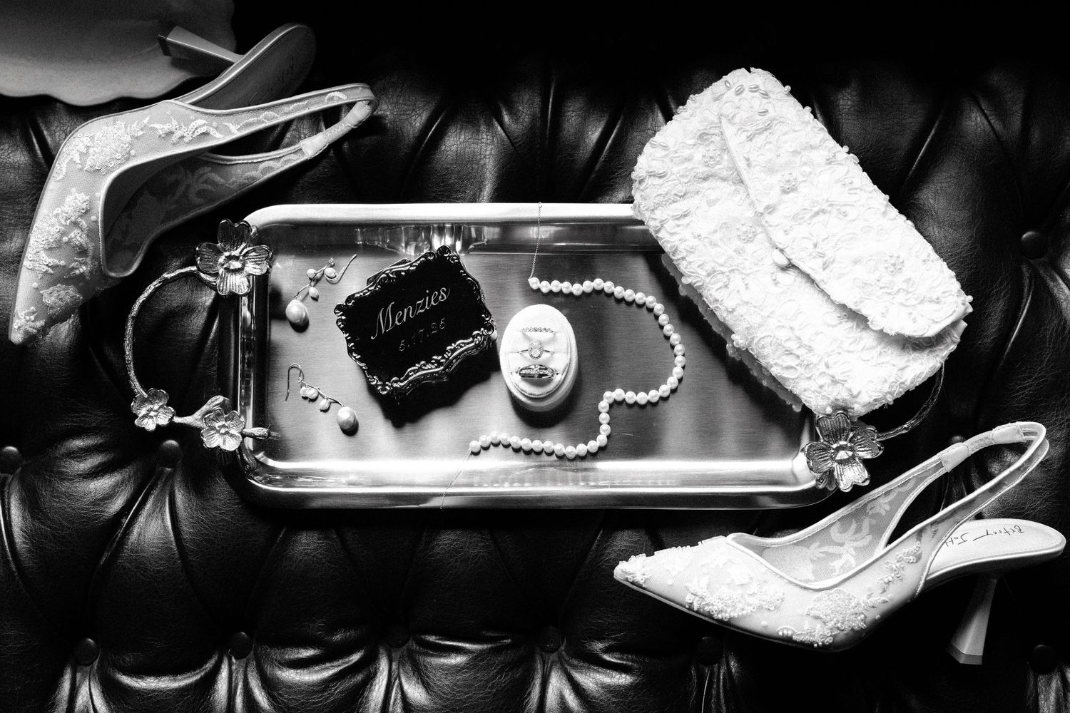 Wedding accessories including white lace heels, jewelry, and a clutch arranged on black leather surface.