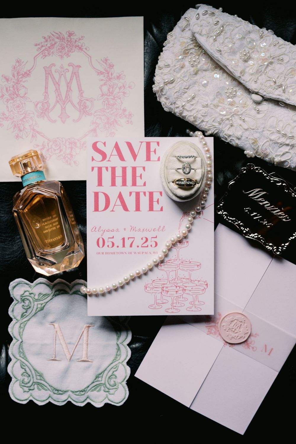 Elegant wedding stationery collection featuring pink save the date cards, pearl necklaces, and delicate accessories on dark surface.