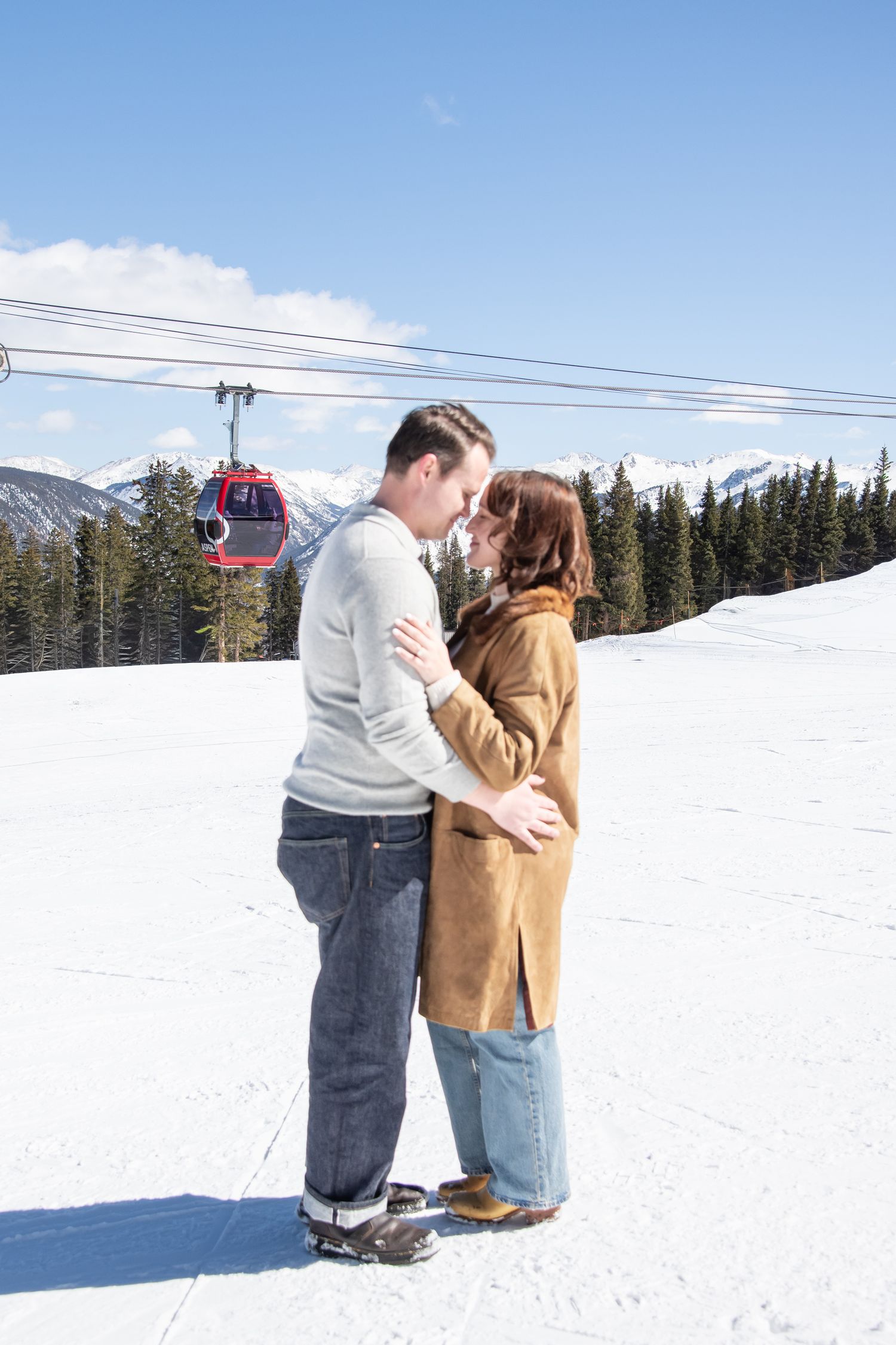 Winter engagement photos with a cute young couple on top of the gondola with a red gondola car and mountains with snow in the background
