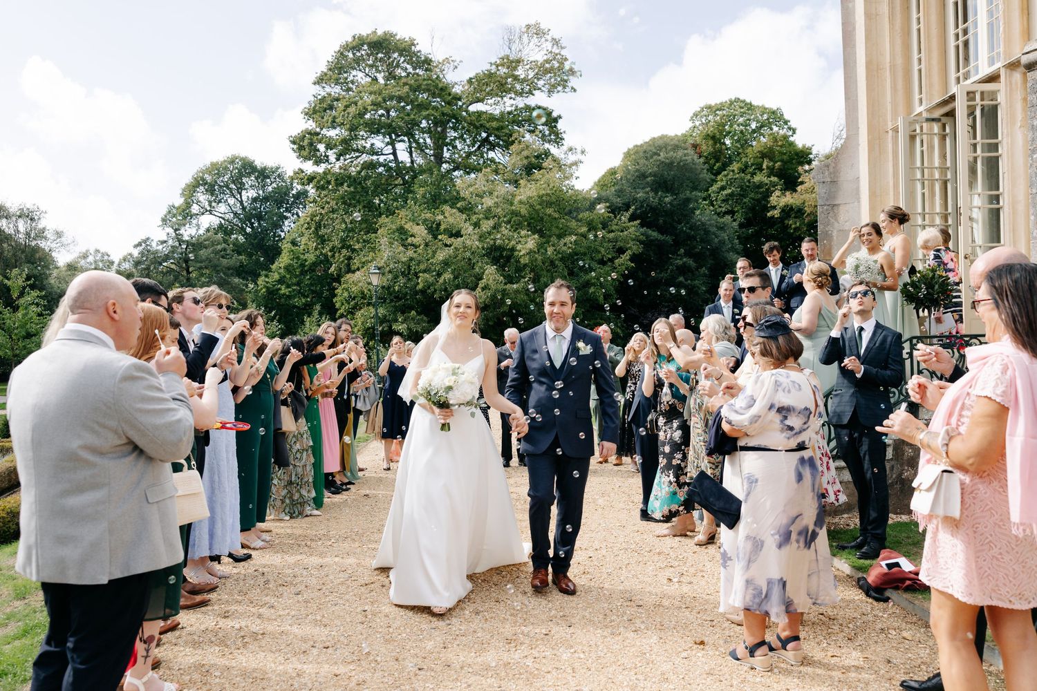 Wedding guests gather on a garden path outside a grand estate during an elegant outdoor celebration.