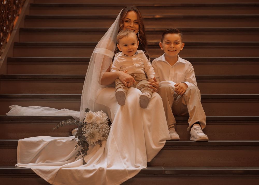 Bride in white gown and veil sits on wooden staircase with two young boys, holding a bouquet of white flowers.