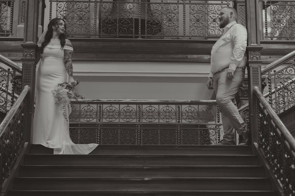 Black and white photo of bride and groom posing on ornate staircase in wedding attire.