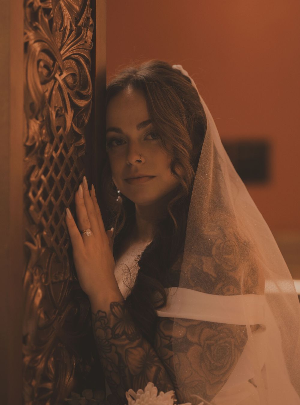 Beautiful tattooed bride in lace veil poses beside ornate carved door at wedding venue.