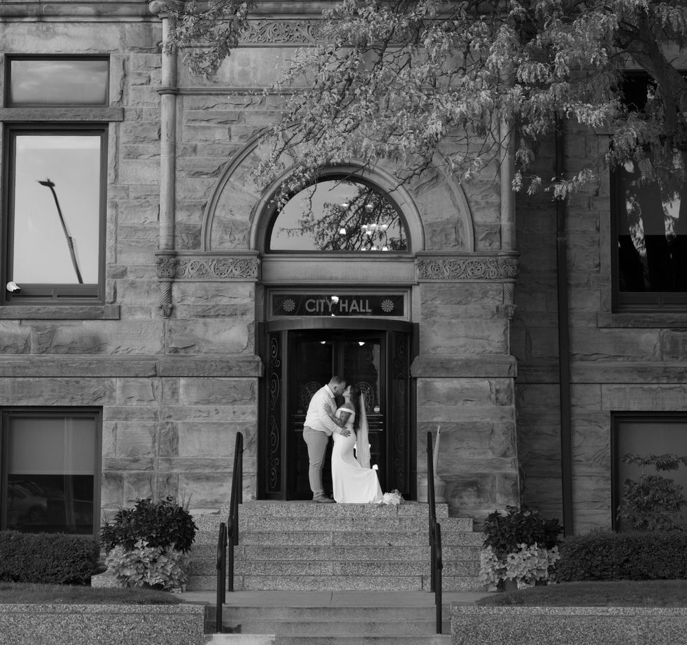 Newlyweds share a kiss on the steps of a historic City Hall in black and white photo.