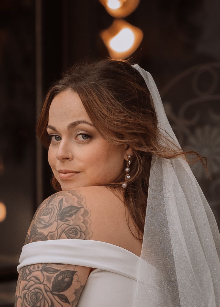 Bride with floral tattoo on shoulder wearing white gown and veil, pearl earrings, soft bridal makeup.