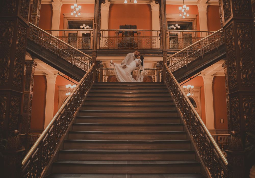 Bride and groom embrace on grand ornate staircase in elegant wedding venue with warm lighting.