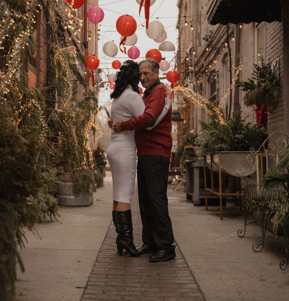 A couple embraces romantically in a decorated alley with red lanterns and twinkling lights overhead.