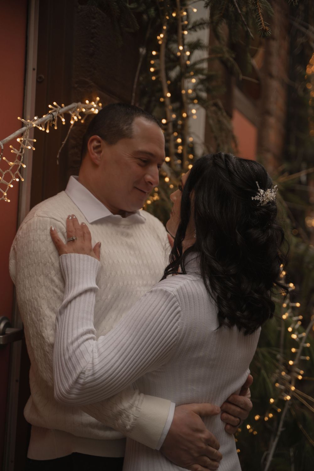 A couple in matching white sweaters embrace romantically outdoors, surrounded by warm fairy lights and evergreen branches.