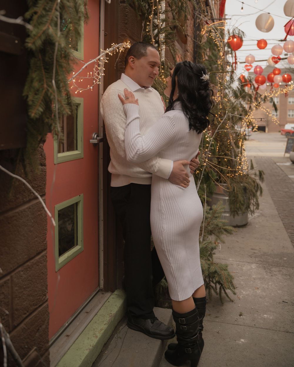 Couple sharing a romantic moment leaning against a red building surrounded by string lights and lanterns.