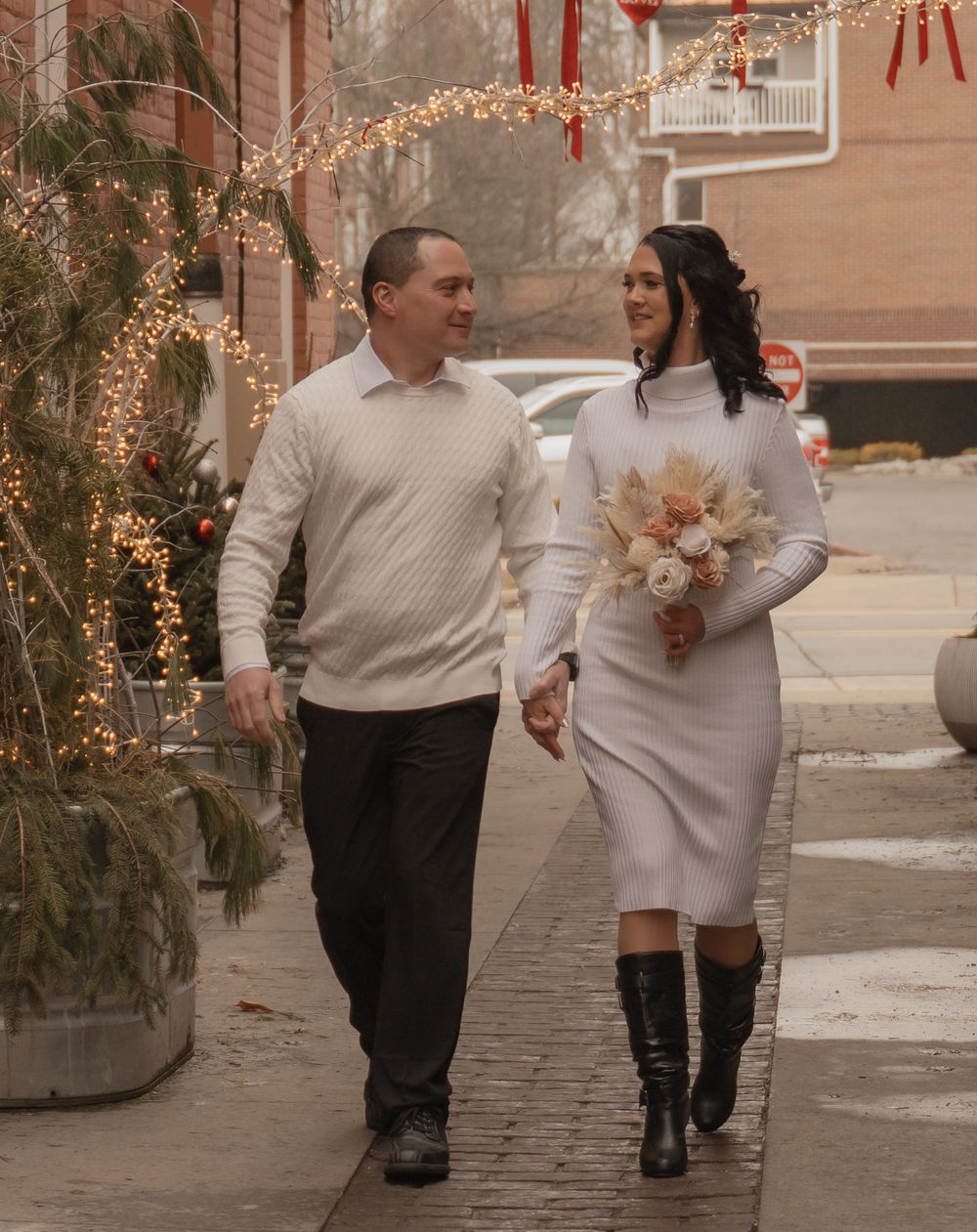 Couple in matching white outfits walking hand in hand on a festive street decorated with string lights.