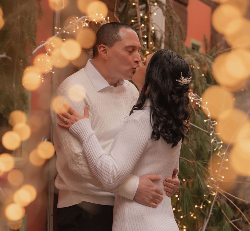A couple shares a tender kiss surrounded by warm bokeh fairy lights and festive greenery.