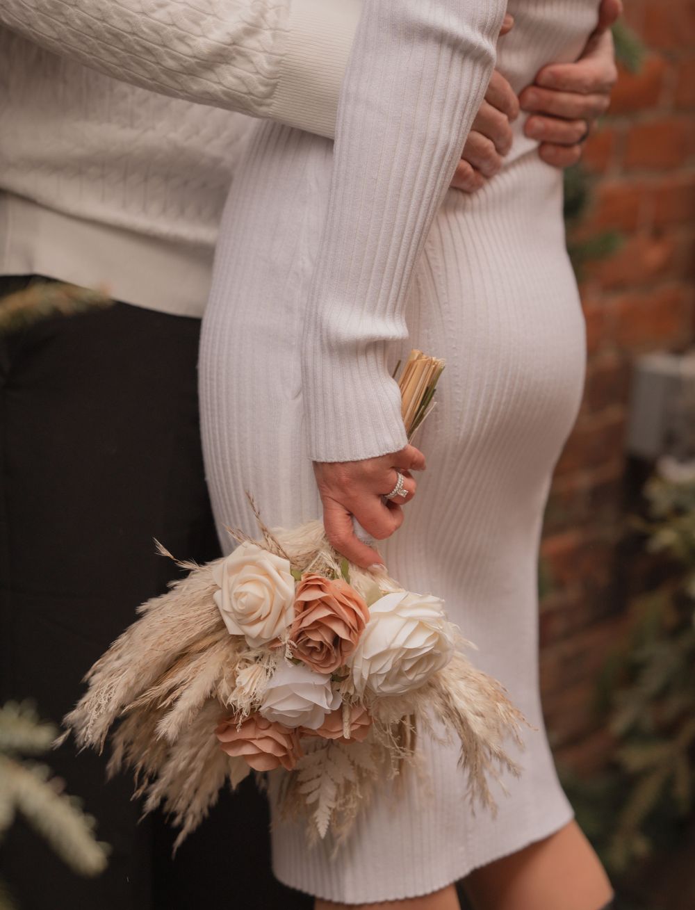 Bride in white ribbed dress holding a bouquet of blush roses and pampas grass while embracing her partner.