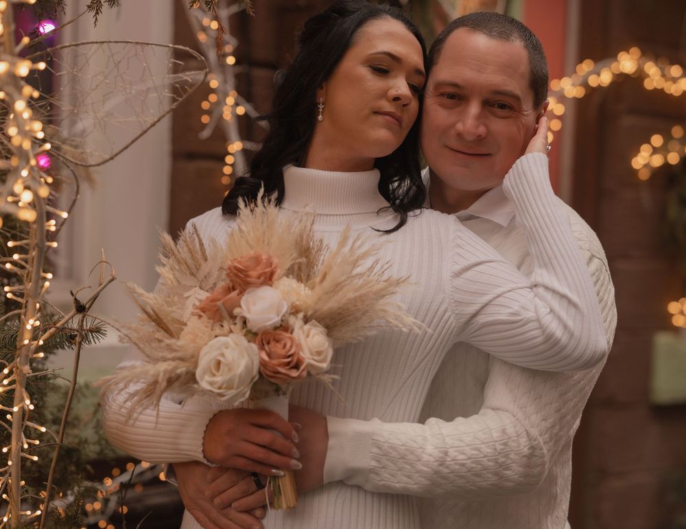 Couple in matching white sweaters embracing near Christmas tree, bride holding dried floral bouquet with blush roses.