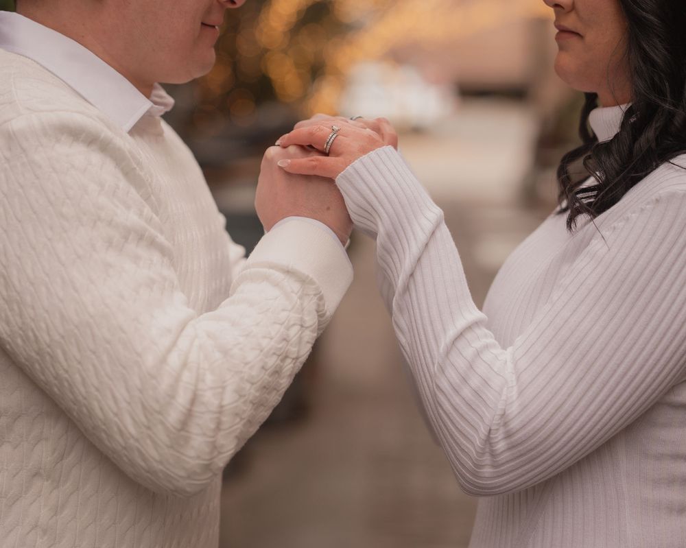 Close-up of a couple holding hands, woman wearing an engagement ring, dressed in white sweaters.