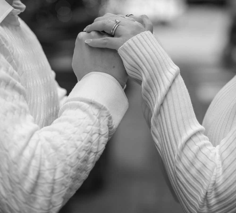 Black and white photo of two people holding hands tenderly, wearing light-colored long-sleeve shirts.