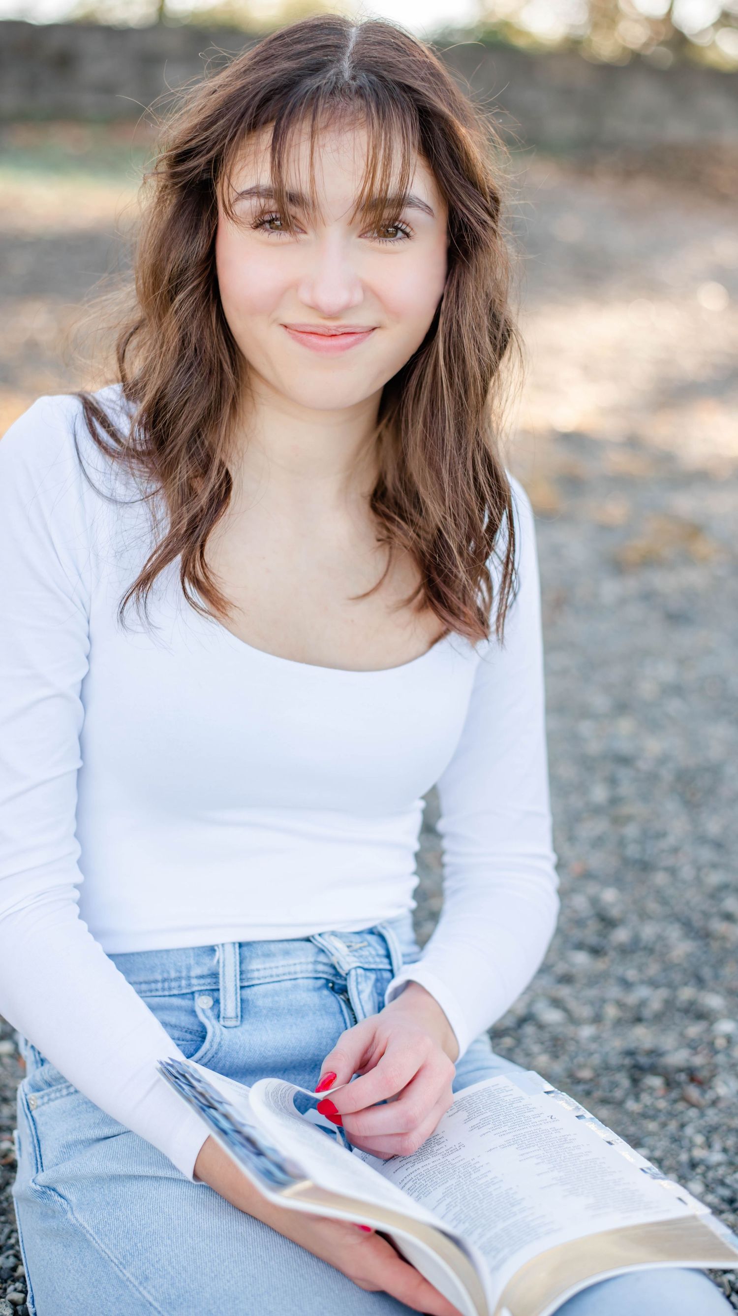 Young woman in white top and jeans sitting outdoors reading a book, smiling at camera.