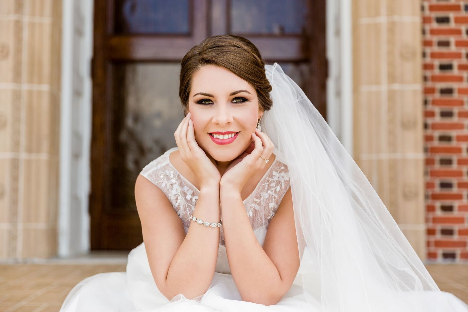 Smiling bride in white lace gown and veil, resting chin on hands, posing outdoors near brick building.