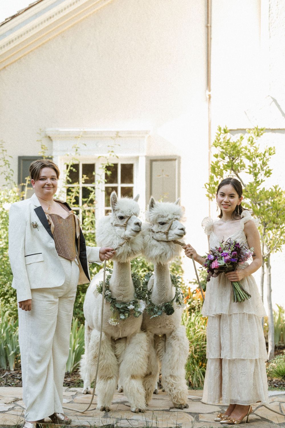 Two brides posing with alpacas nuzzling together with floral necklaces at The Memorial House SLC Utah alpaca rental
