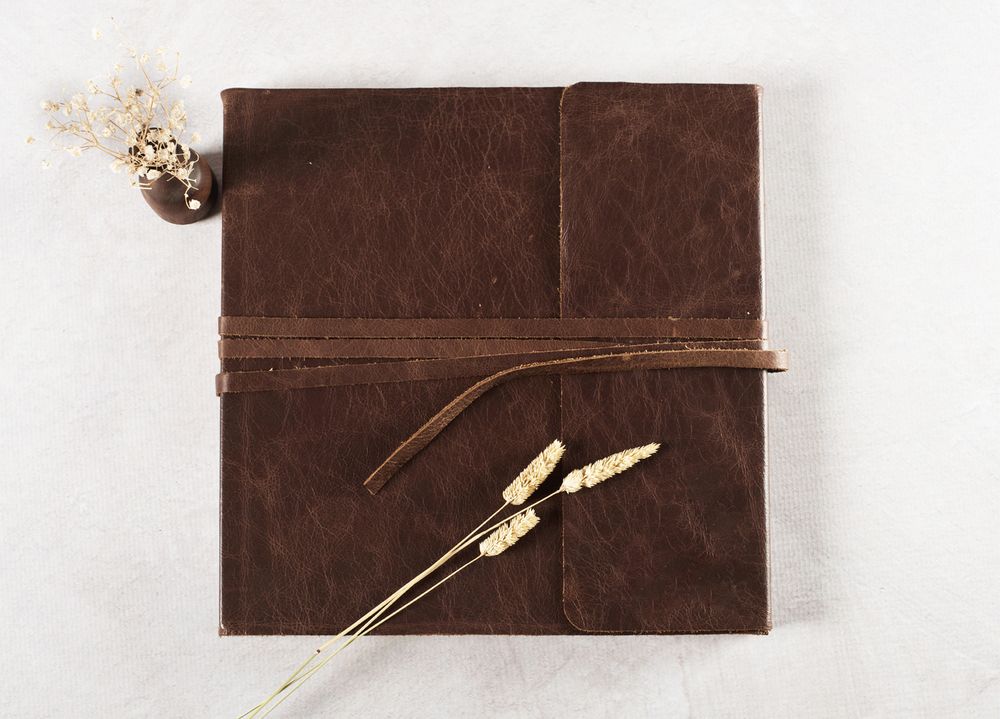 Closed brown leather journal tied with strap, styled with dried wheat and small floral sprig on white background.