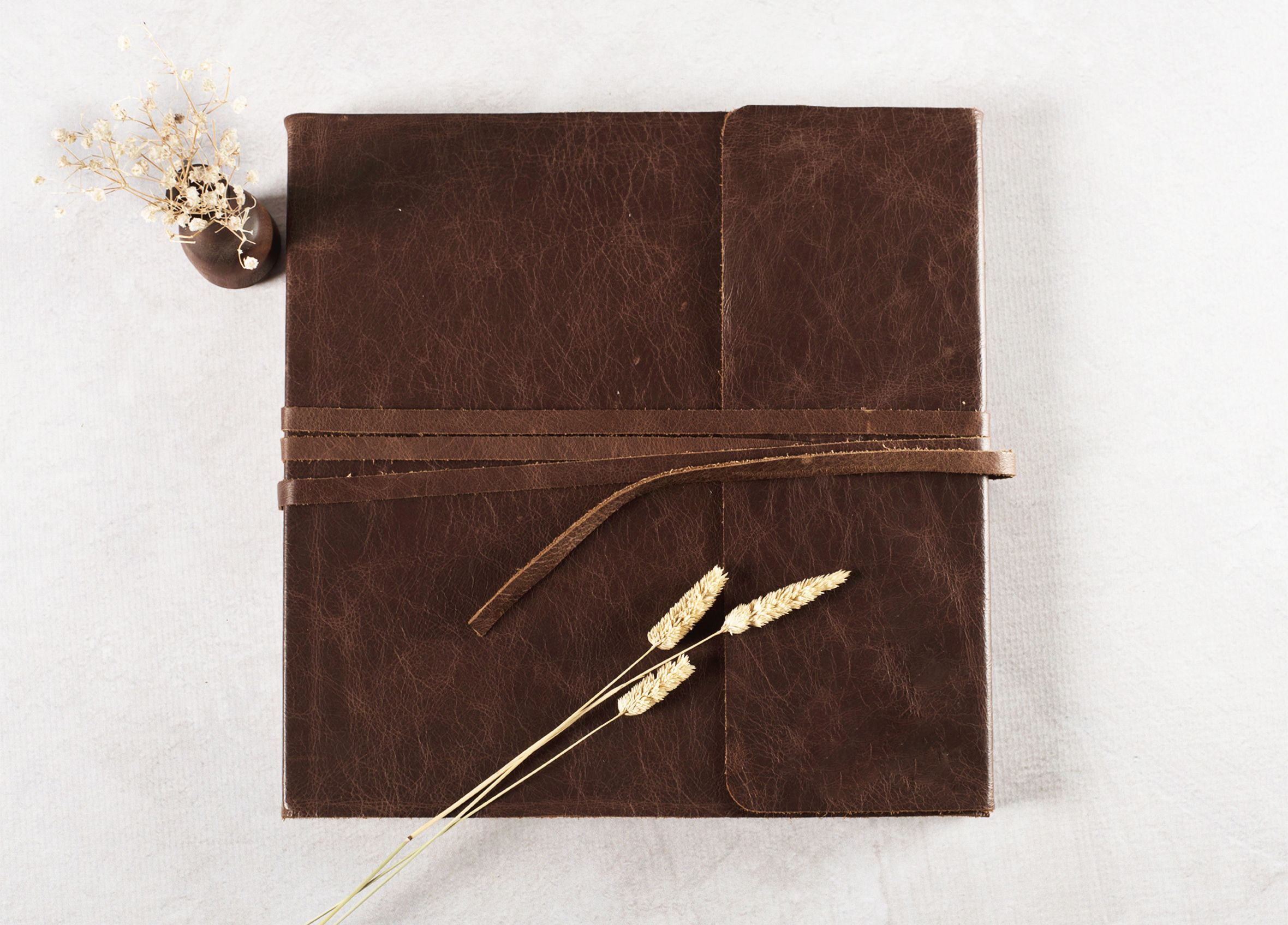 Closed brown leather journal tied with strap, styled with dried wheat and small floral sprig on white background.