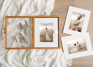 Wooden photo box with dried flowers beside framed beach wedding portraits on white linen backdrop.