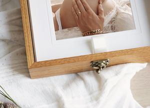 Wooden keepsake box with photo frame lid displaying a bridal wrist wearing a gold bracelet, placed on white fabric.