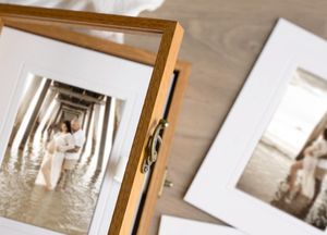 Two framed wedding photos displayed on a wooden surface, showcasing a couple standing under a pier in shallow water.
