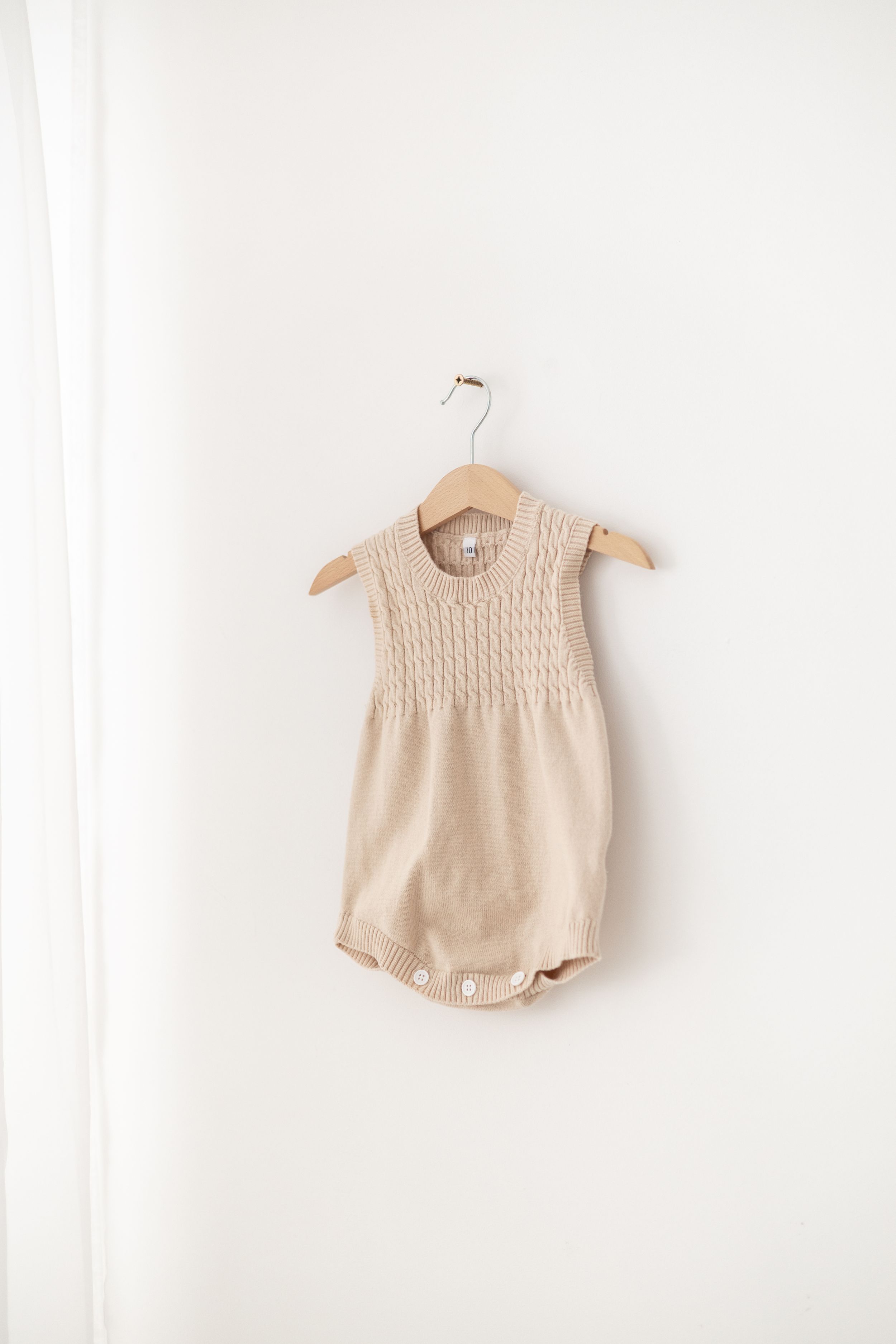 Beige knit baby romper with cable pattern detail hanging on wooden hanger against white wall.
