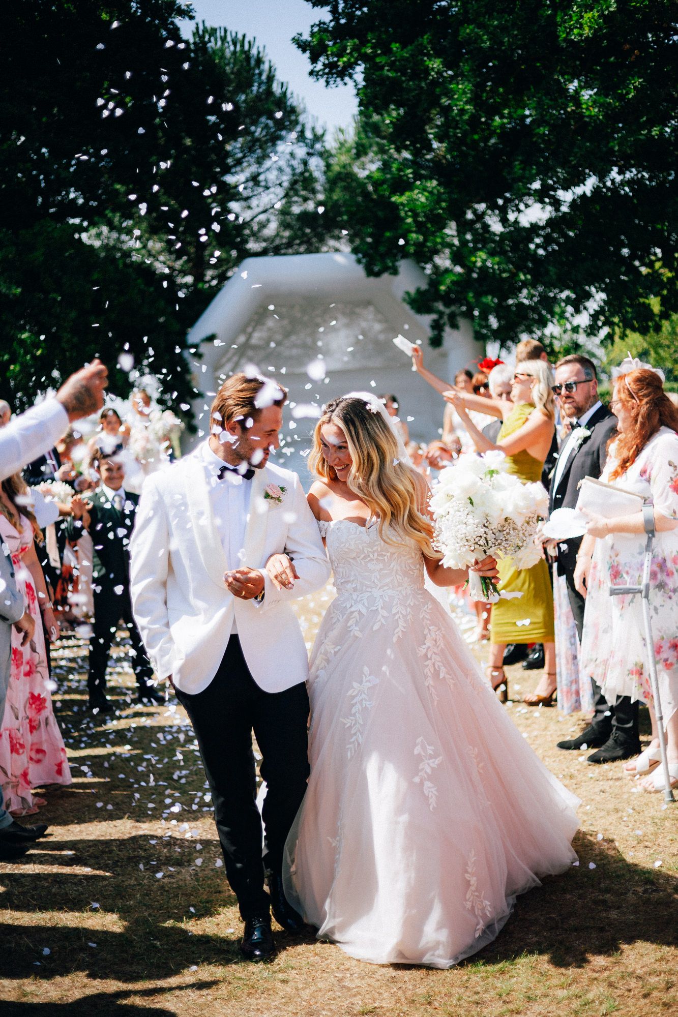 Bride and groom walk through confetti shower after outdoor wedding ceremony, bride in lace ballgown holding white bouquet.