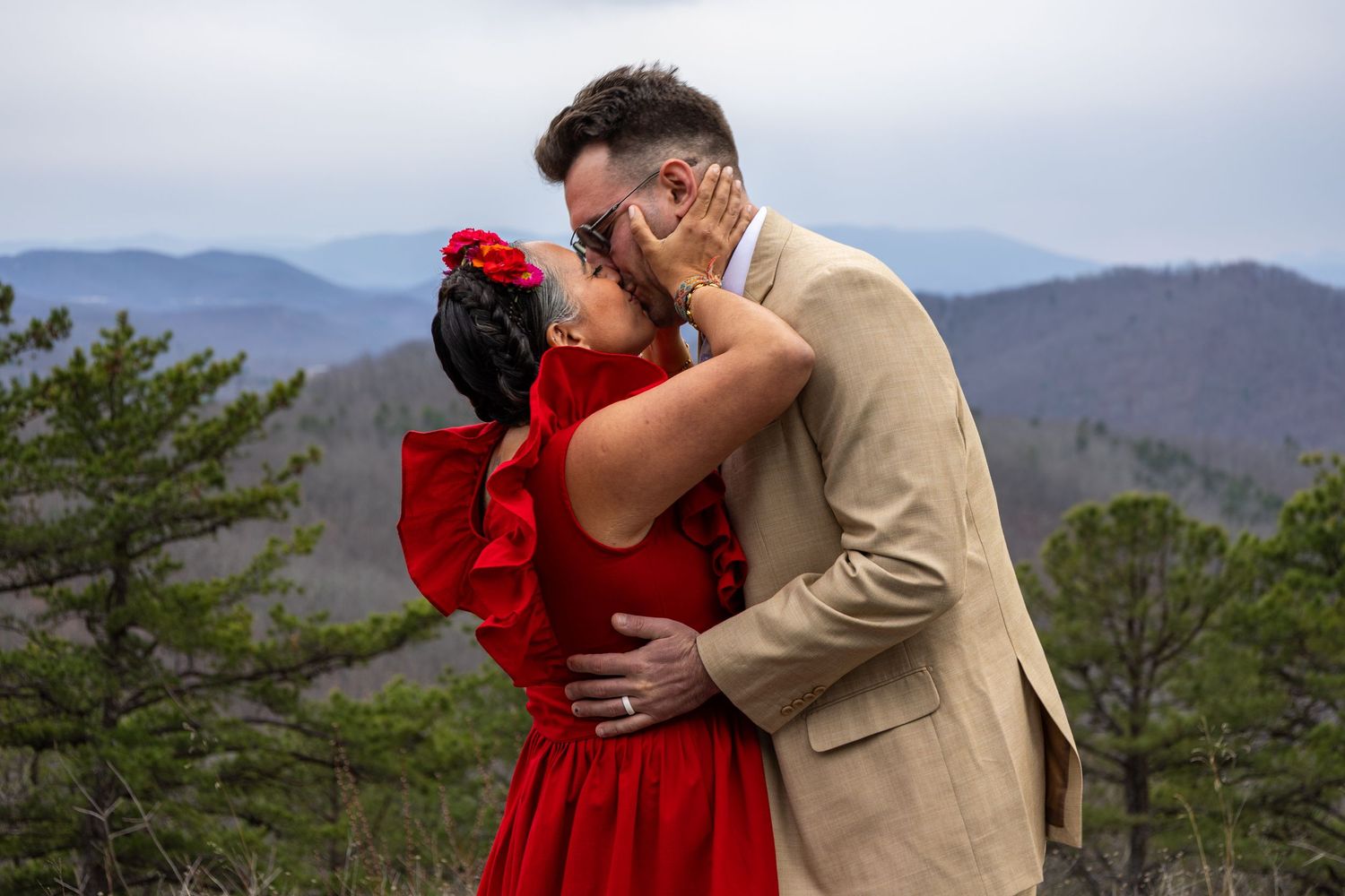 Romantic couple kissing on scenic mountain overlook, woman in vibrant red dress.