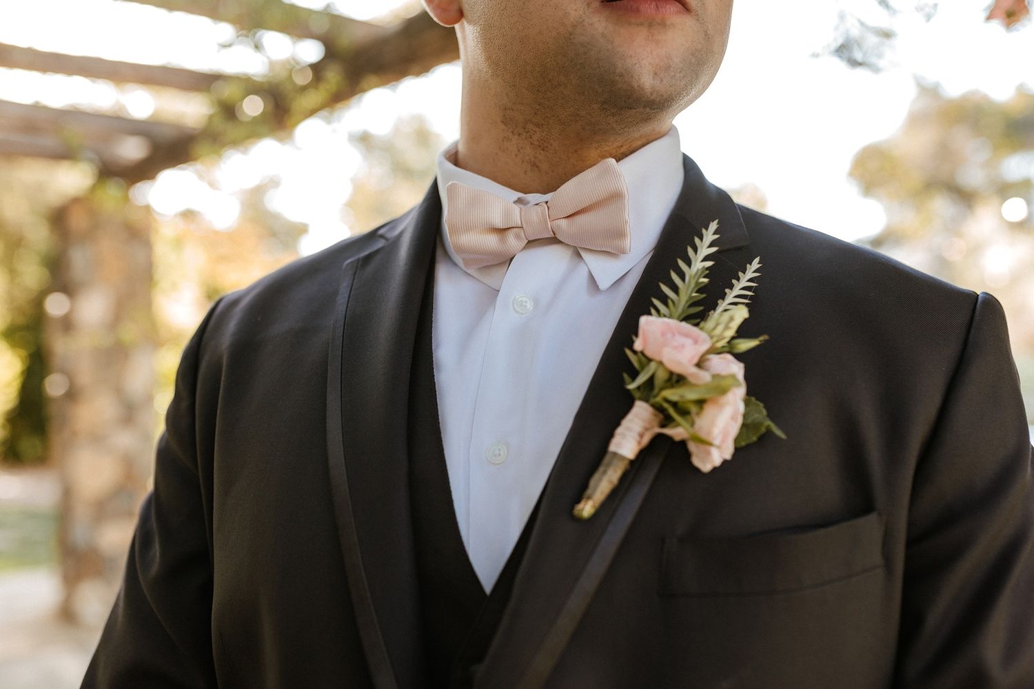 Groom in black tuxedo with blush pink bow tie and floral boutonniere at wedding.