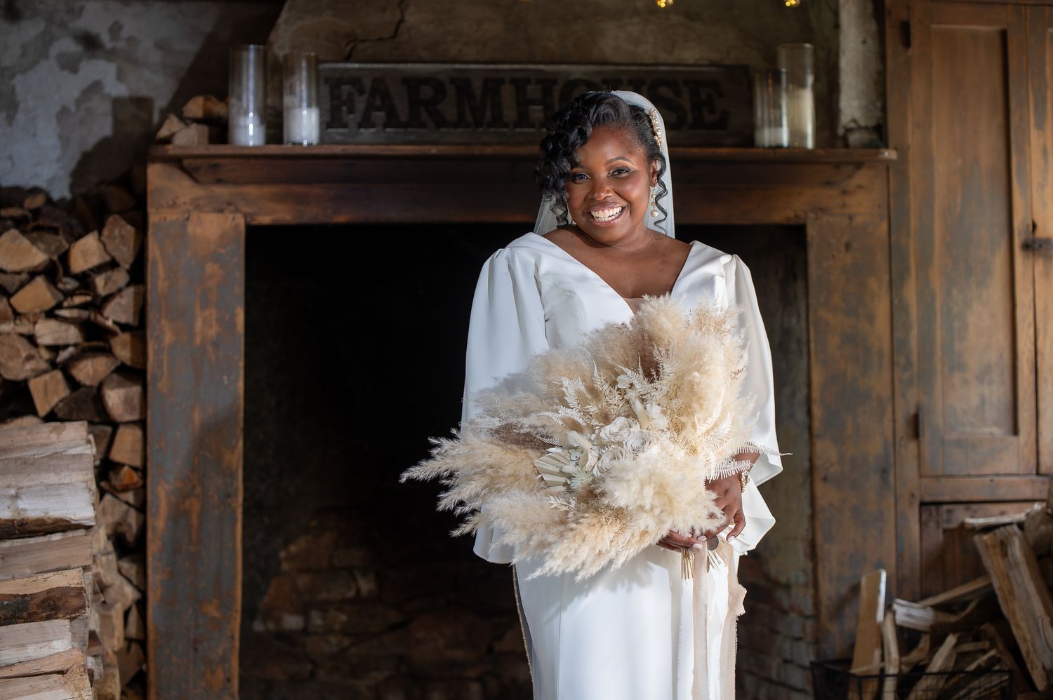 Woman in white dress holding dried pampas grass bouquet, smiling in front of rustic farmhouse doorway.