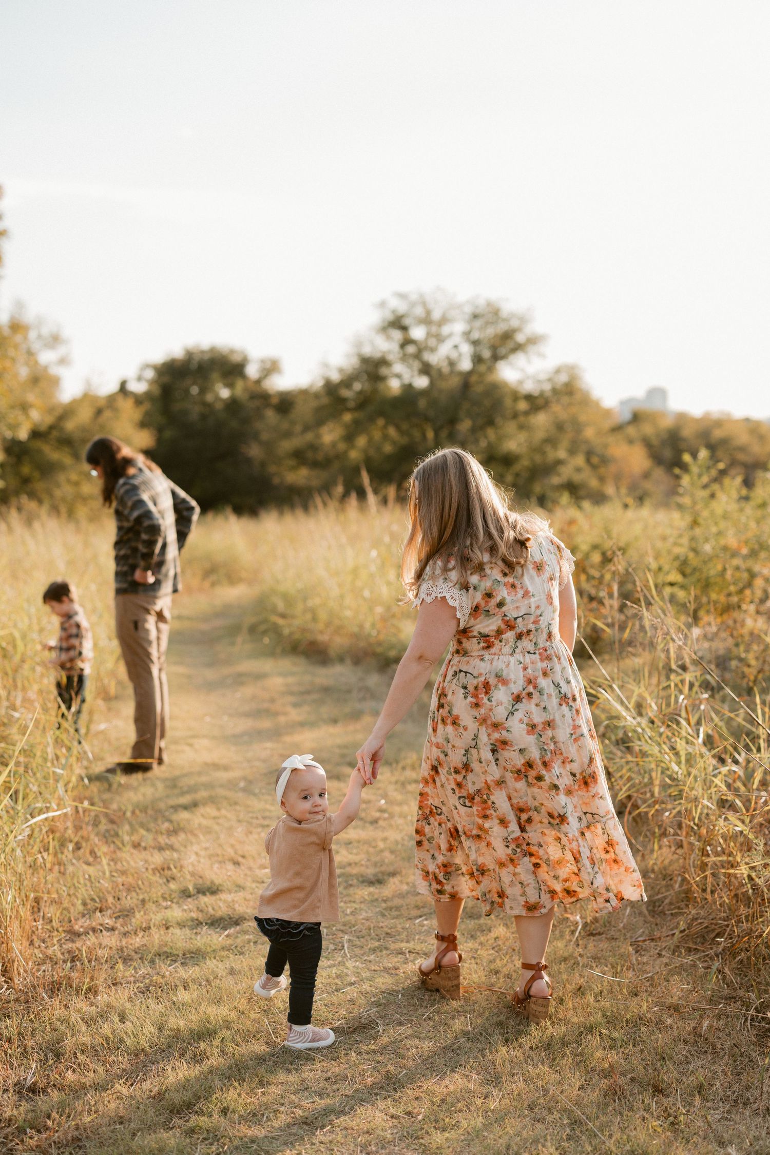 Family walking on a golden field path in autumn, mother in floral dress holding toddler's hand, father with child in background.