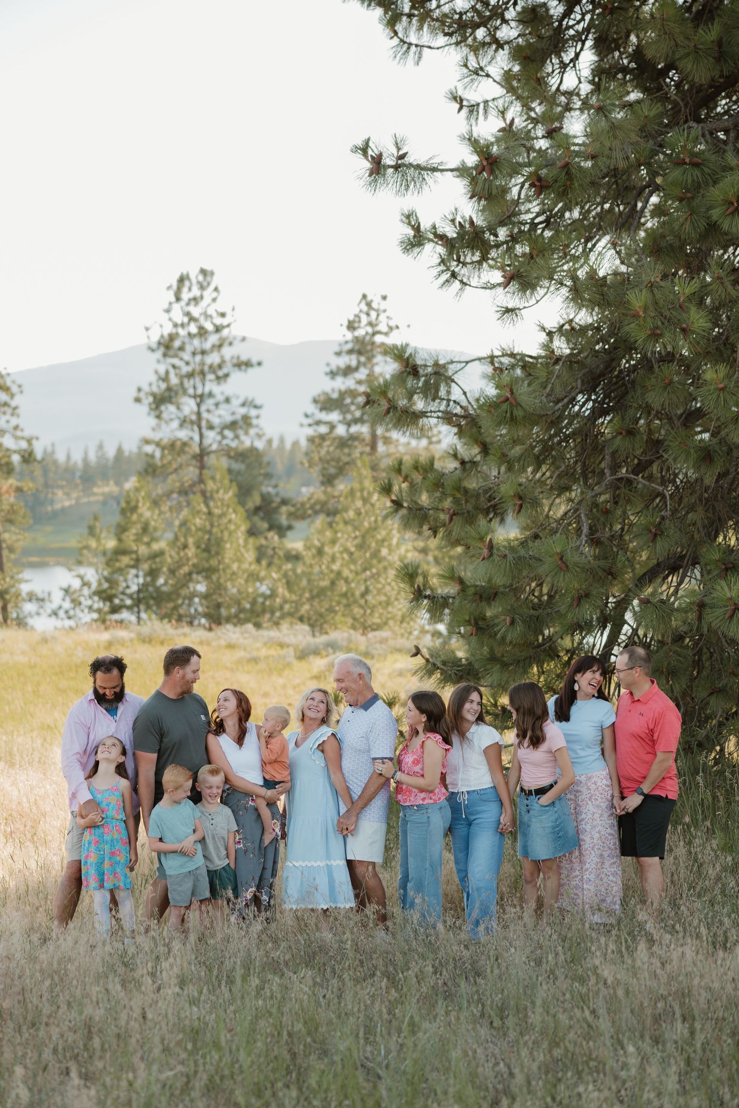Large family group portrait outdoors in golden meadow with pine trees and mountain lake backdrop during sunset.