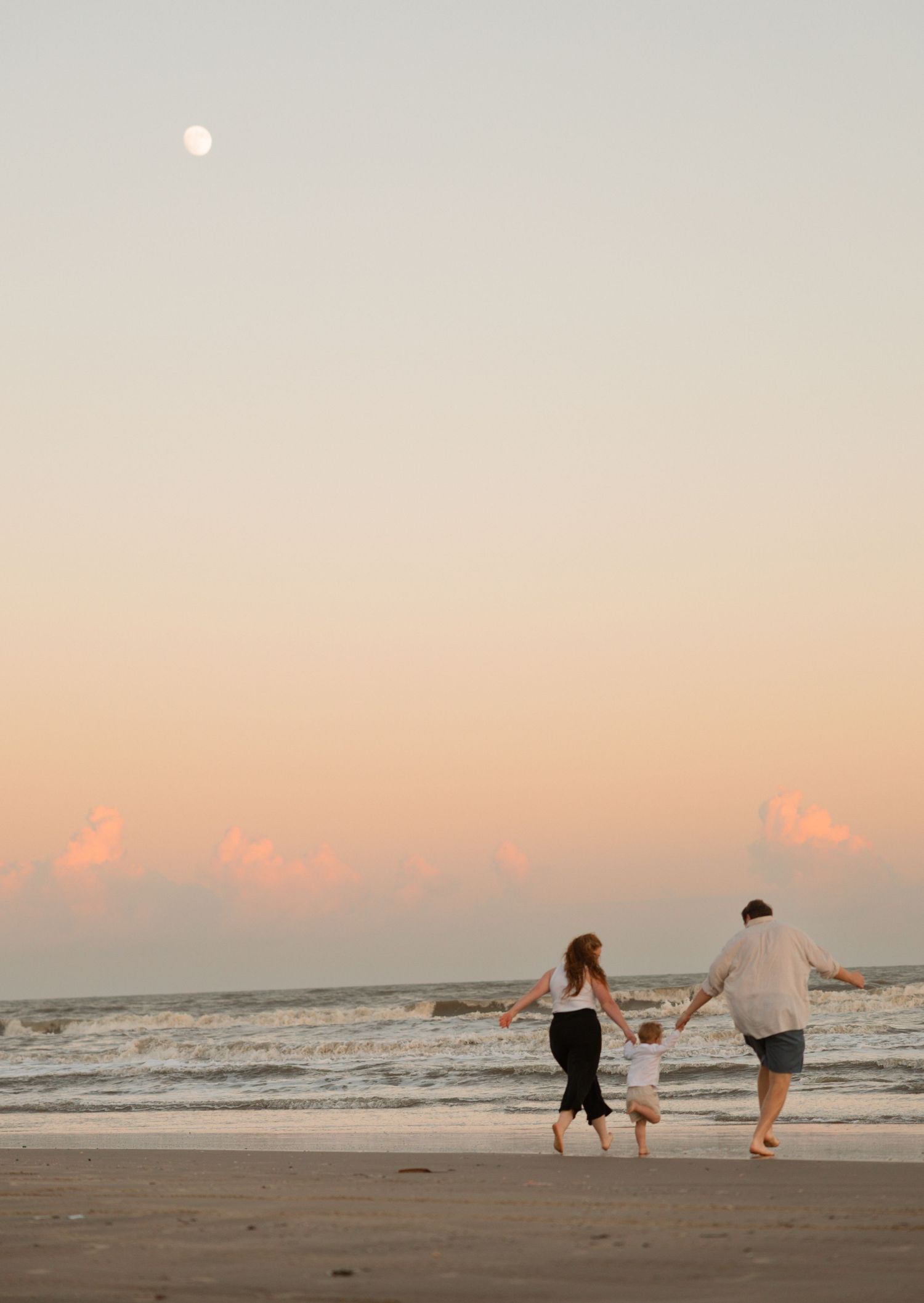 Family of three holding hands walking on beach at sunset with moon visible in pastel sky.