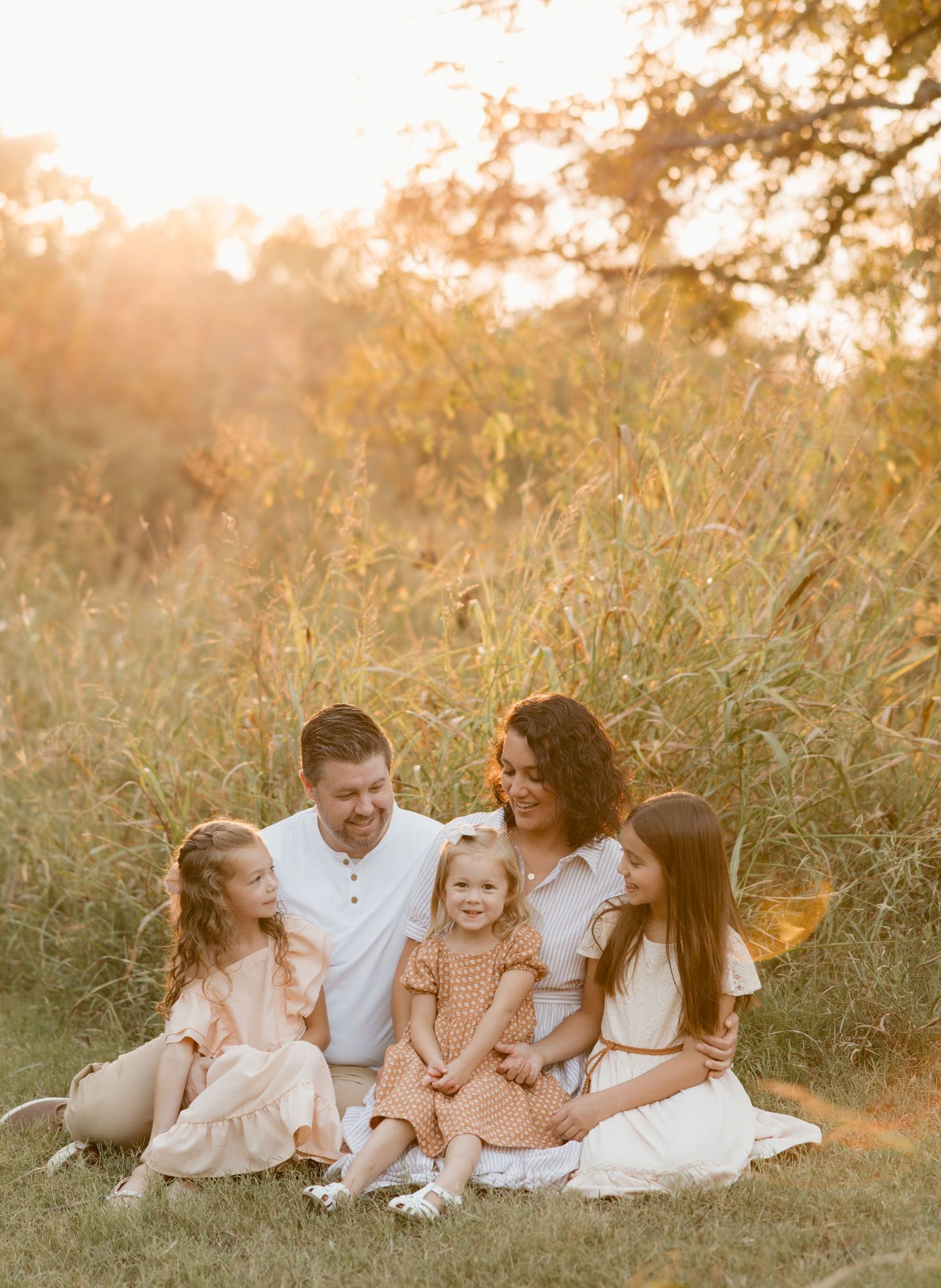 Family of five sitting together in golden hour sunlight, smiling warmly in a grassy field with tall reeds behind them.