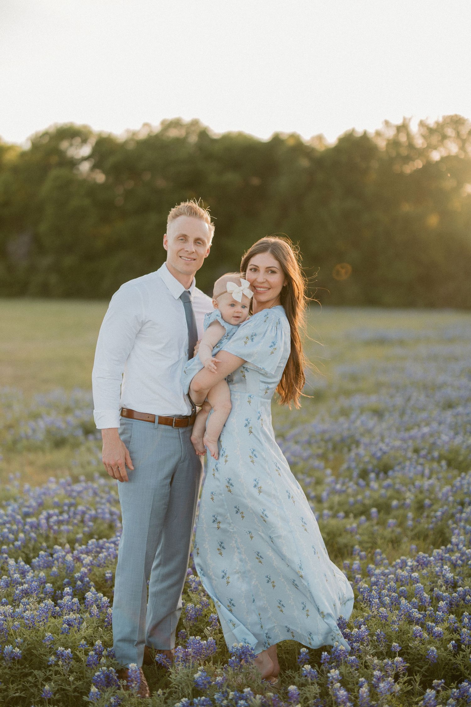 Family portrait of couple holding baby in bluebonnet field at golden hour, woman in floral dress, man in gray suit.