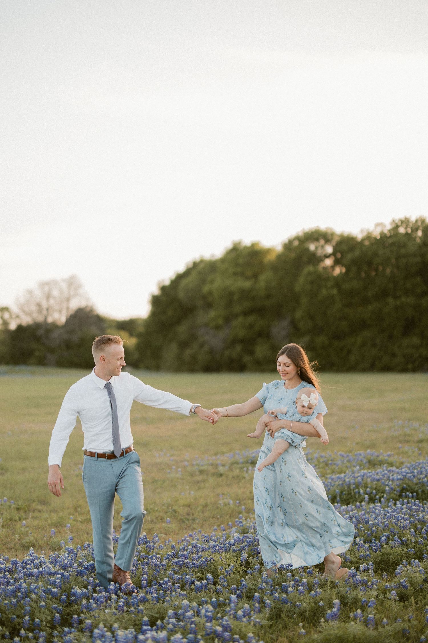 Couple holding hands in a bluebonnet field at golden hour, woman in floral dress holding baby, man in white shirt.