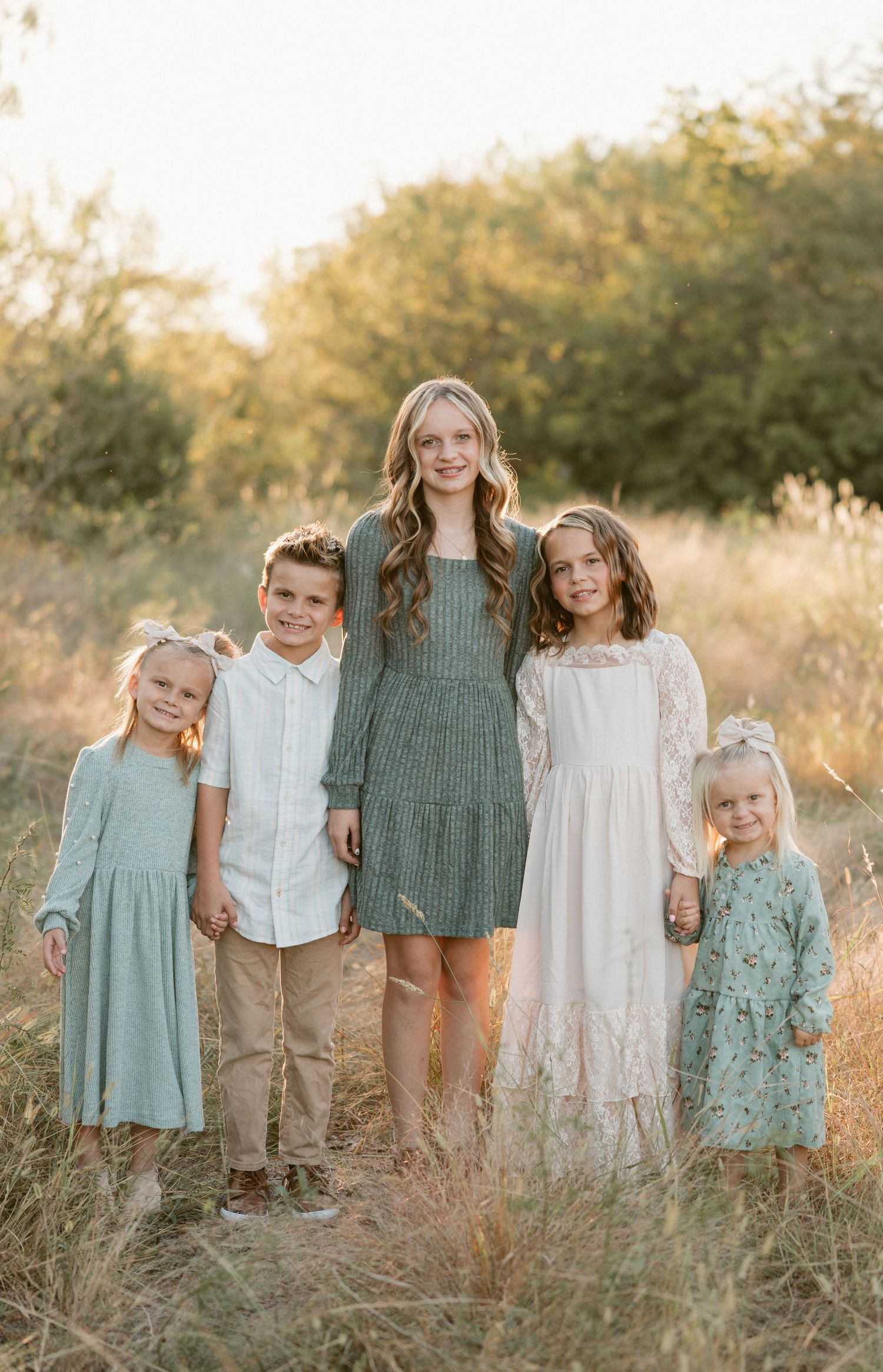 Five children smiling together outdoors in a sunlit field, dressed in coordinating sage green and white outfits.