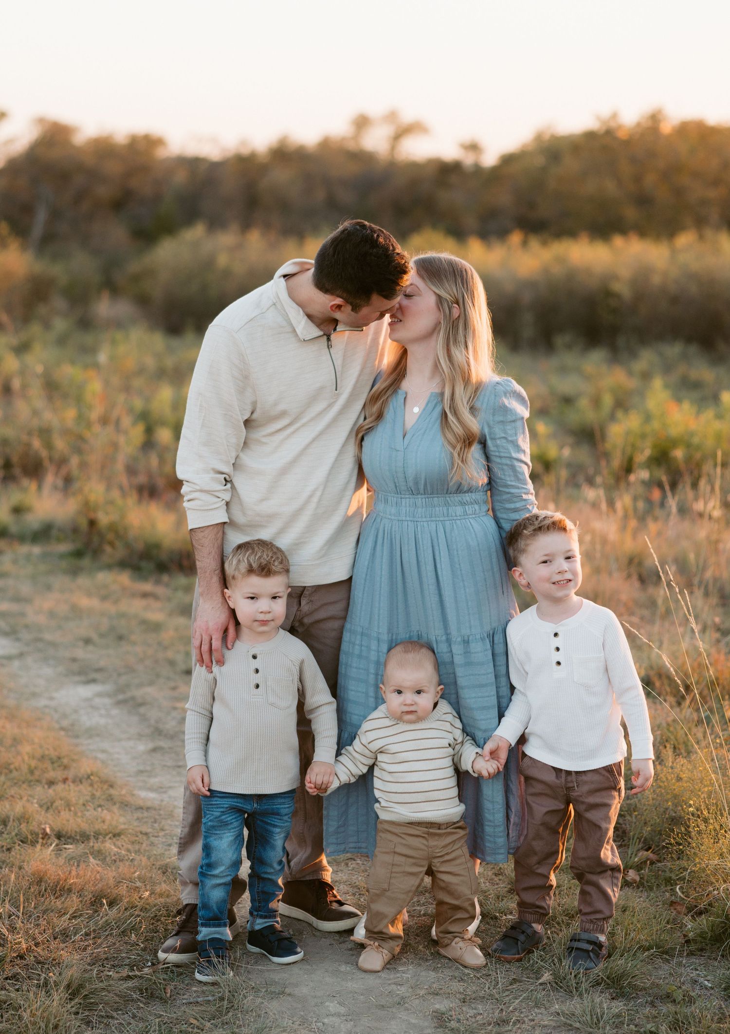 A young family with two toddlers and a baby poses for a golden hour portrait on a sunlit dirt path.