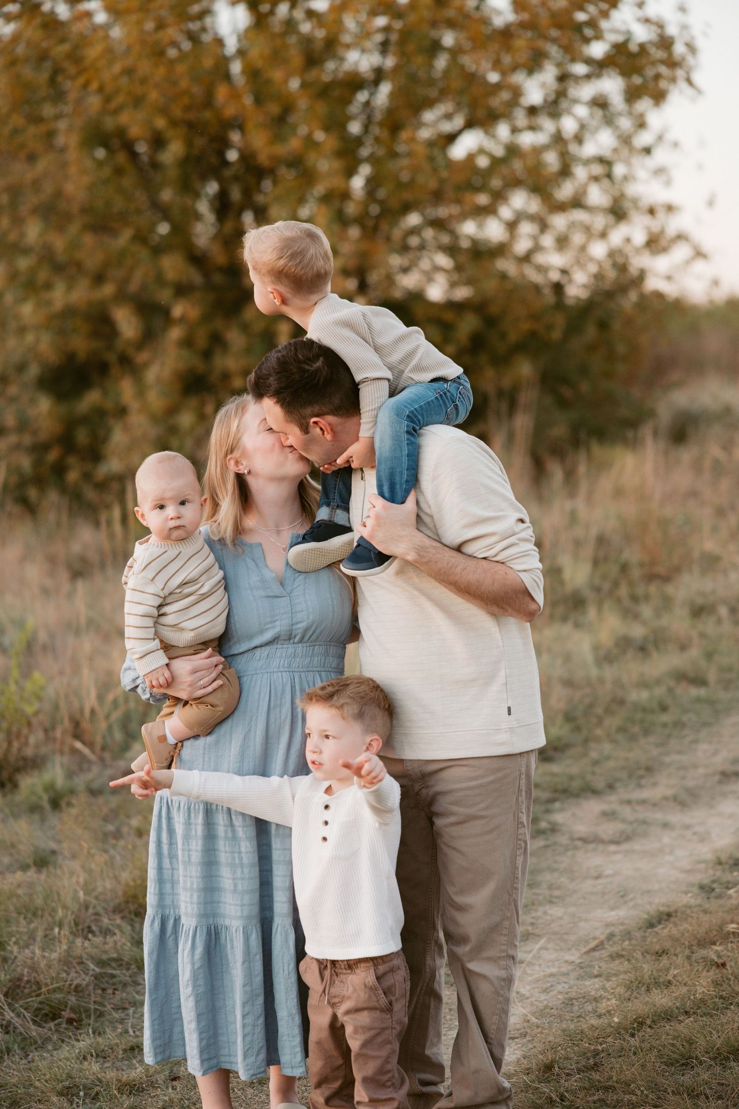 A happy family of five poses outdoors at golden hour, parents kissing while holding three young children in a field.