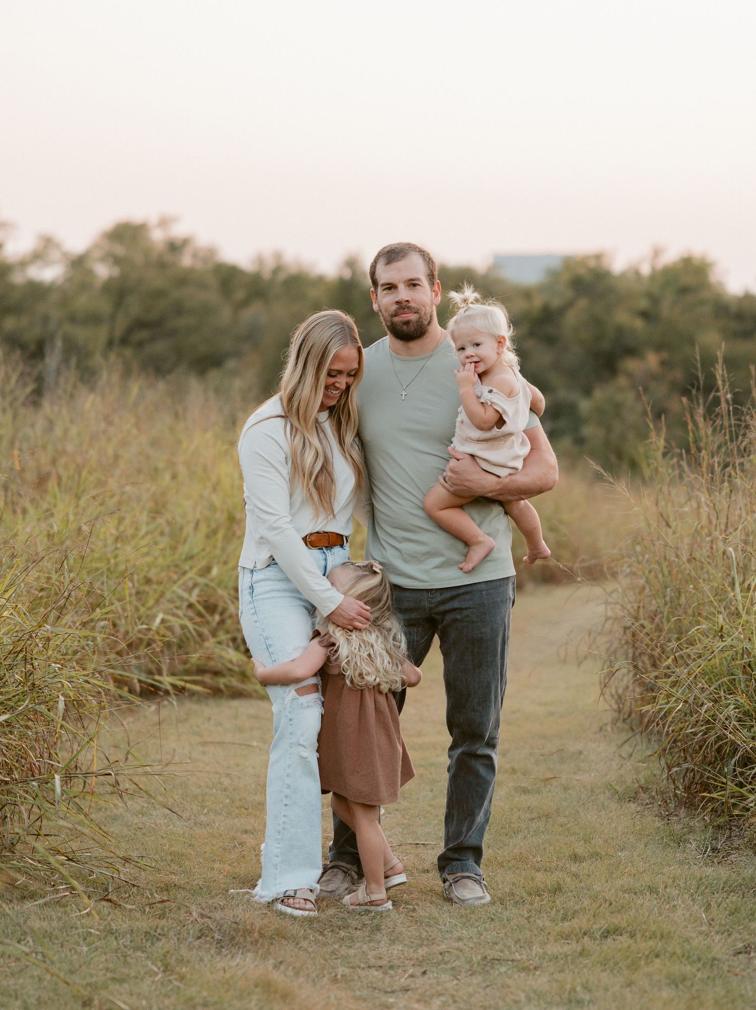 A young family poses on a grassy path at sunset, with man holding baby girl and woman beside them.