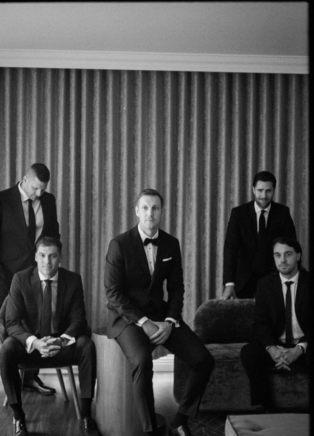 Five men in formal black suits pose together in a stylish black and white wedding party portrait indoors.